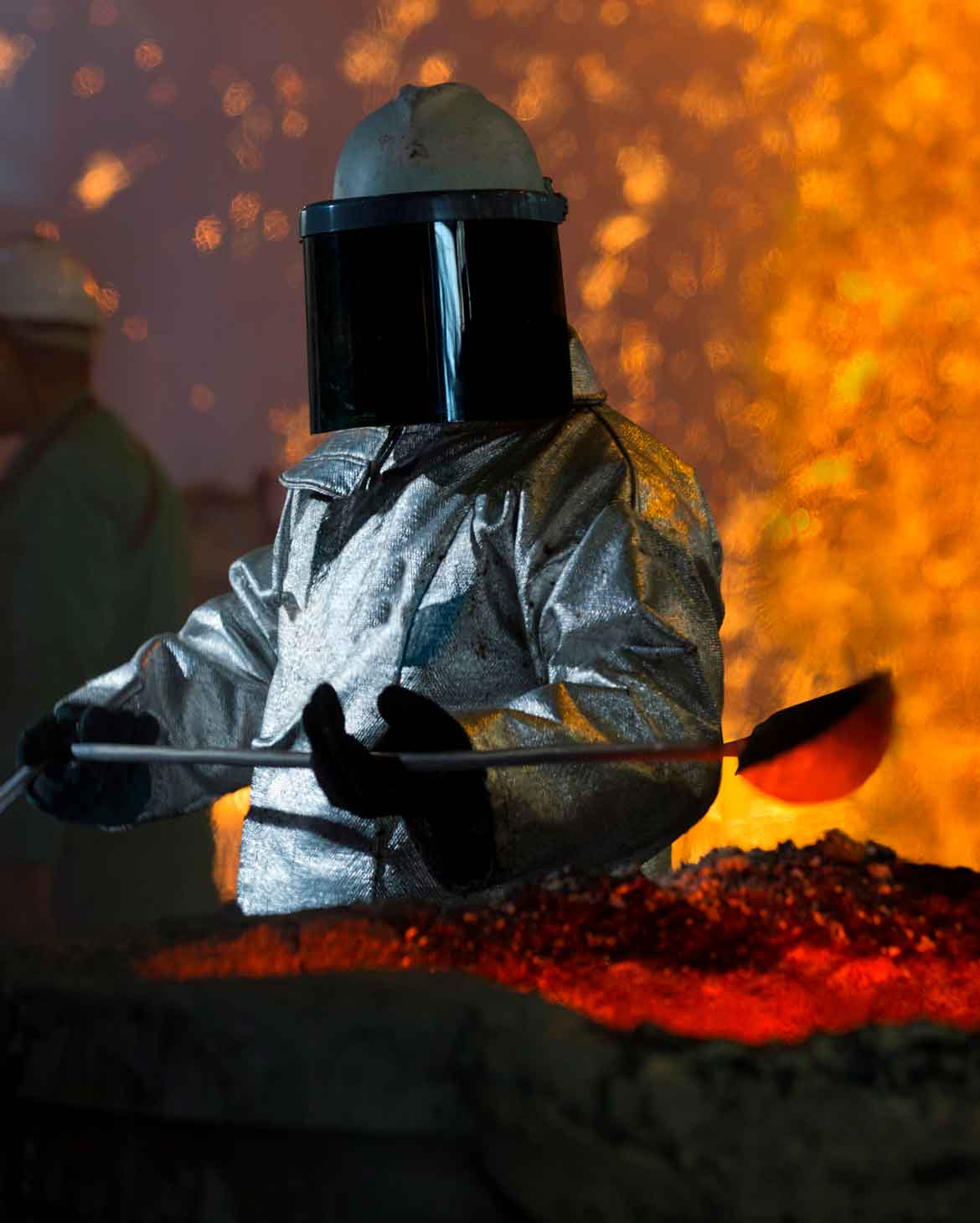 trabalhador industrial vestindo conjunto aluminizado completo com capacete e protetor facial escuro operando ferramenta perto de material fundido incandescente e faíscas em ambiente de fundição