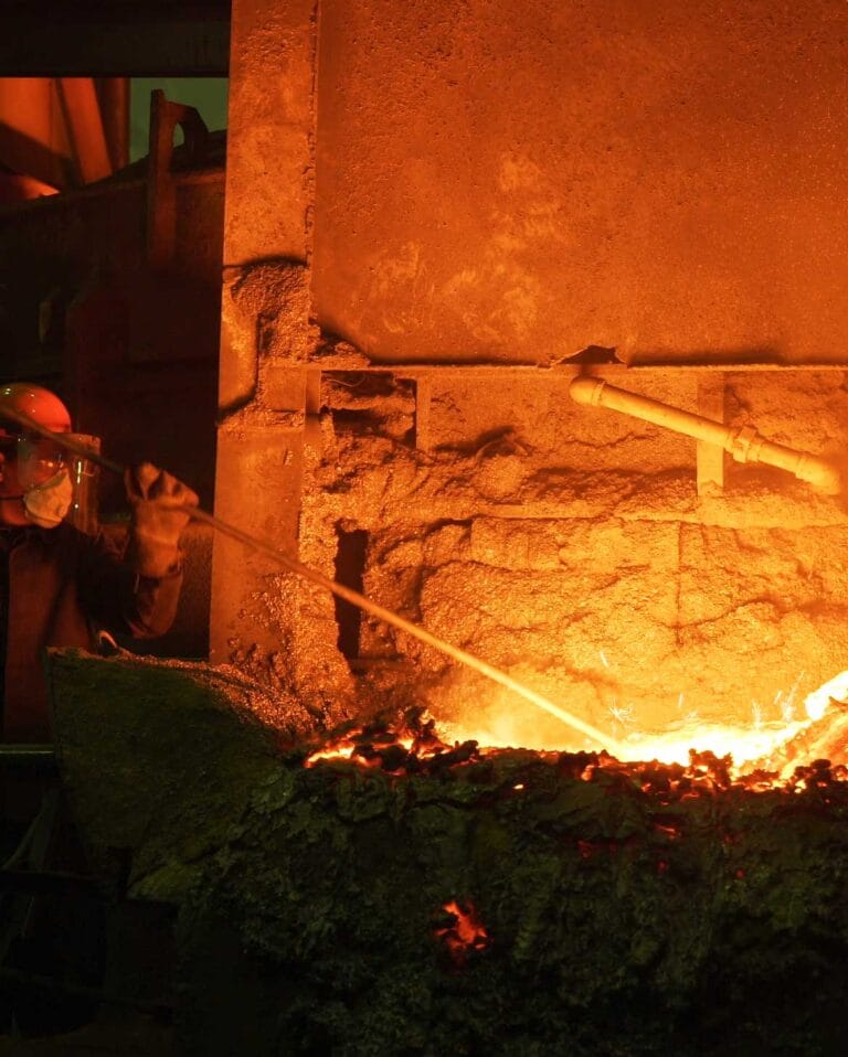 Trabalhador em metalúrgica utilizando EPIs para fundição, como protetor facial e respirador, para manipular com segurança o metal incandescente em um forno industrial.