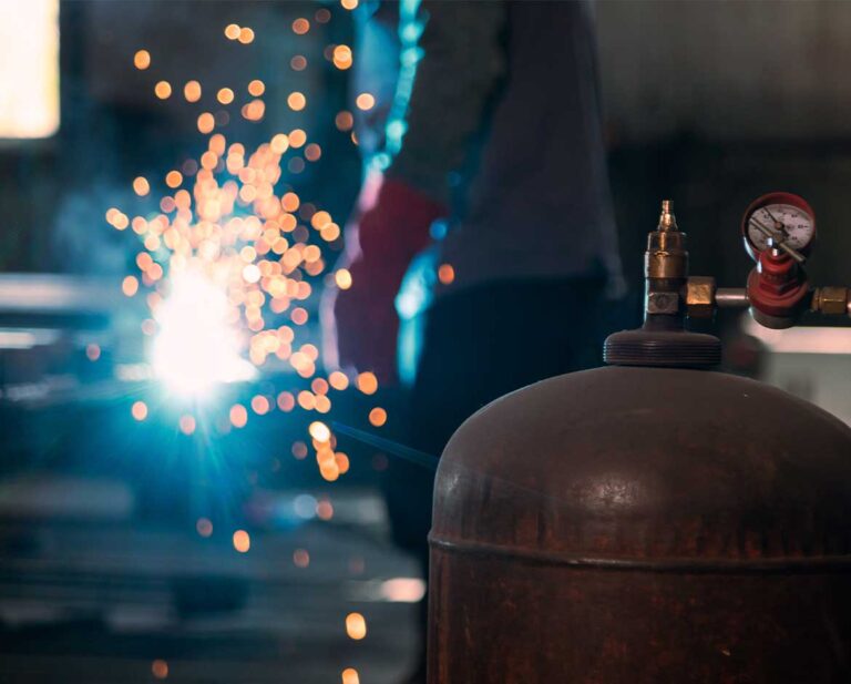 Cilindro de gás em primeiro plano com soldador ao fundo realizando trabalho a quente com faíscas