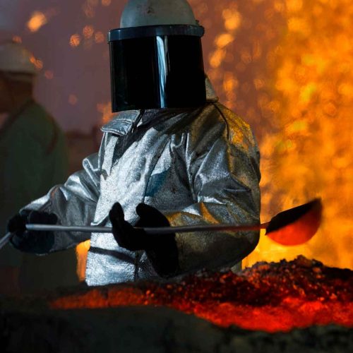 trabalhador industrial vestindo conjunto aluminizado completo com capacete e protetor facial escuro operando ferramenta perto de material fundido incandescente e faíscas em ambiente de fundição