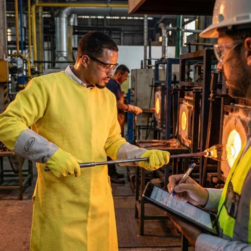 trabalhador com epis de aramida em operação de alta temperatura na indústria do vidro acompanhado por profissional de sst