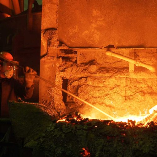 Trabalhador em metalúrgica utilizando EPIs para fundição, como protetor facial e respirador, para manipular com segurança o metal incandescente em um forno industrial.