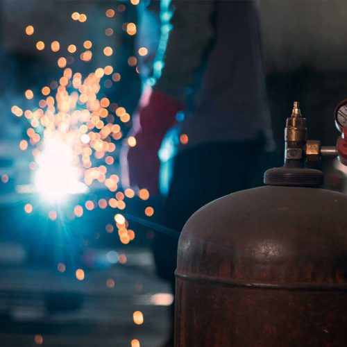 Cilindro de gás em primeiro plano com soldador ao fundo realizando trabalho a quente com faíscas